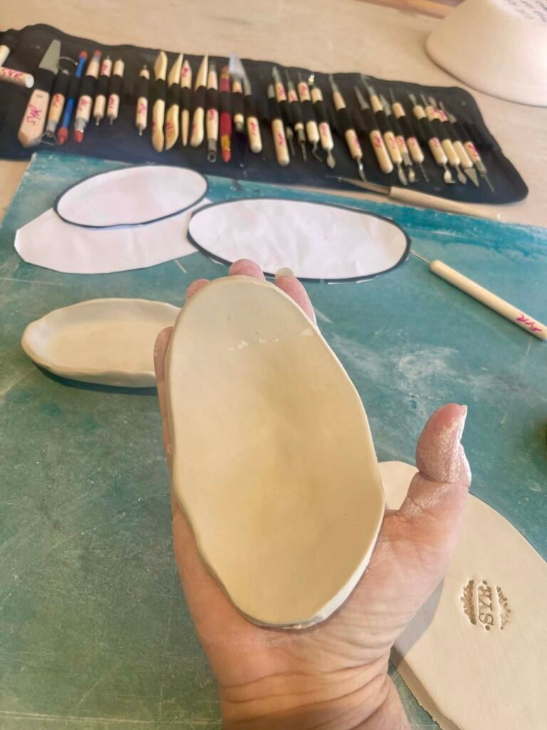 Hands shaping a smooth clay pebble at a workbench, with pottery tools alongside — the clay component of the Still Cracked program